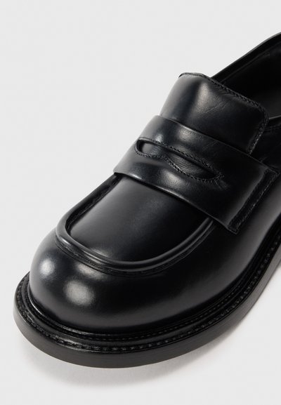Svart läderloafer med rund tå, med en bred rem över framfoten och synlig sömnad längs sulan. Slät yta.