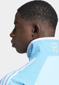 Ljusblå idrottsjacka med vita accenter, hög krage och logotyp på ryggen. Har tre vita ränder på ärmarna och strukturerad tyg.