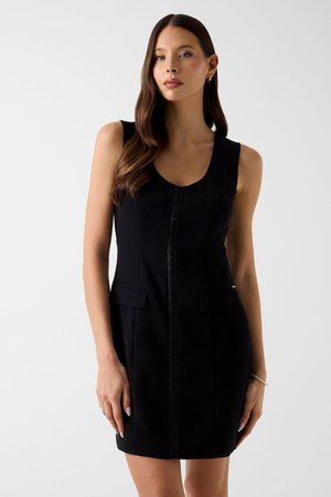 Donna con lunghi capelli castani che indossa un vestito nero senza maniche con bottoni sul davanti e tasche con pattina, in piedi davanti a uno sfondo chiaro e uniforme.