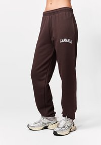 Brązowe spodnie dresowe z elastycznym pasem, z haftowanym napisem "LAMANIA" w kolorze białym. Gładka faktura i zwężane nogawki.
