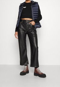 Marineblaue Steppweste, schwarzer, taillierter Pullover mit einem kleinen Design, hoch geschnittene schwarze Kunstlederhose und braune Schnürstiefel bis zum Knöchel.