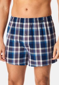 Des boxers à carreaux bleus et blancs présentent une taille élastique, une fermeture à bouton et une texture en coton douce. Les motifs incluent des carrés bleus foncés et clairs.