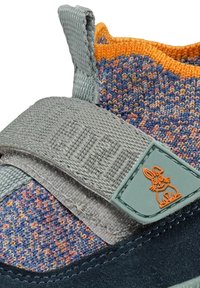 Gros plan sur une chaussure en tricot multicolore pour enfants avec une bordure orange, une bride grise à scratch et un logo lapin sur un patch vert menthe.