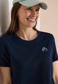 Mujer sonriente que lleva una gorra verde claro y una camiseta azul marino con "BE KIND" y un logo de una palmera en el pecho.