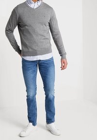 Suéter gris de cuello en V sobre una camisa de cuadros, combinado con jeans azules y zapatillas blancas. El atuendo presenta un corte clásico y un sutil detalle de logo.