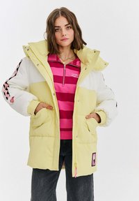 Pufi bunda v žltej a bielej kordovej látke, s predným zipsom, nadrozmernou kapucňou, dvoma prednými vreckami a ružovými akcentmi na rukávoch.