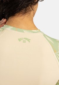 Camisa de surf de manga larga en beige claro con detalles en estampado tropical verde, que presenta un logotipo texturizado y costuras planas en la parte posterior.