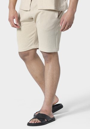 Hombre con pantalones cortos beige texturizados y sandalias negras tipo chancla, de pie con un pie ligeramente levantado sobre un fondo blanco.
