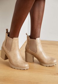 Bottines beiges à cheville avec des panneaux latéraux élastiques, en cuir lisse, à talon bloc et semelle robuste, dotées d'une languette pour un enfilage facile.