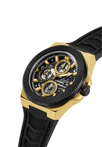 Montre Guess noire et dorée avec un bracelet en caoutchouc noir texturé, des accents dorés angulaires et plusieurs cadrans sur un cadran à motif sombre.