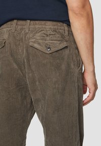 Brązowe spodnie z kordurowej tkaniny o prążkowanej fakturze, posiadające dwie tylne kieszenie z klapkami i guzikiem. Elastyczny pas dla regulowanego dopasowania.