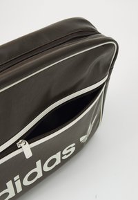 Bruine leren duffeltas met witte accenten, heeft een afgeronde vorm, een opvallende rits en het woord "adidas" in het wit gedrukt.