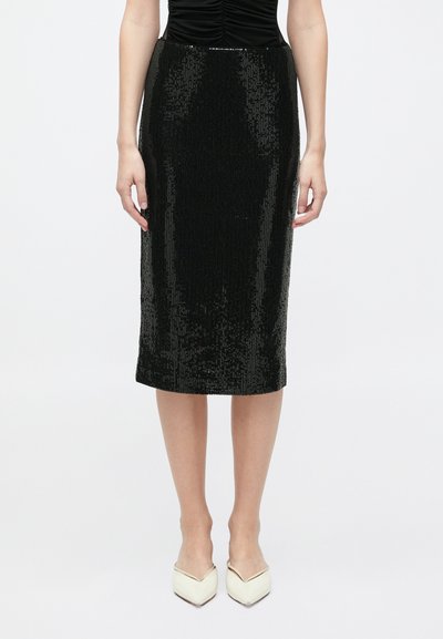 Mujer vestida con una falda lápiz negra de lentejuelas a la altura de la rodilla y unos tacones blancos de punta, posando contra un fondo blanco liso.