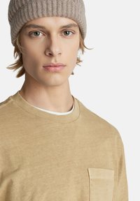 Světlý hnědý crewneck svetr s přední kapsou, vyrobený z měkké, texturované látky. Kombinován se světle béžovou žebrovanou pletenou čepicí.