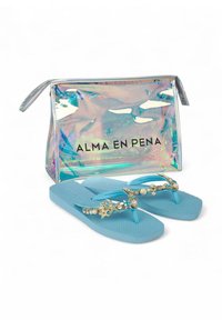 Chanclas azules con suelas texturizadas presentan una correa decorativa adornada con conchas y perlas. Acompañadas de un bolso transparente e iridiscente.
