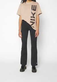 Ljusbeige T-shirt med svart Nike-logotyp på framsidan, i kombination med svarta utsvängda byxor och svarta snörkängor, placerade mot en vit bakgrund.