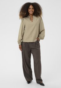 Világos olívazöld blúz V-nyakkal és redőzött ujjakkal, sötétbarna framtás szerű nadrággal és fekete hegyes orrú cipővel párosítva.