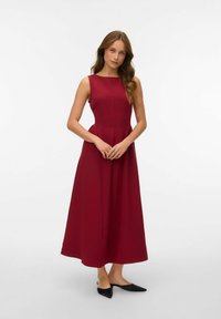 Burgundijas bezpalaku maksi kleita ar pieguļošu augšdaļu un izplestu svārku, kas izceļas ar vienkāršu dizainu un gludu tekstūru. Melni apavi papildina apģērbu.