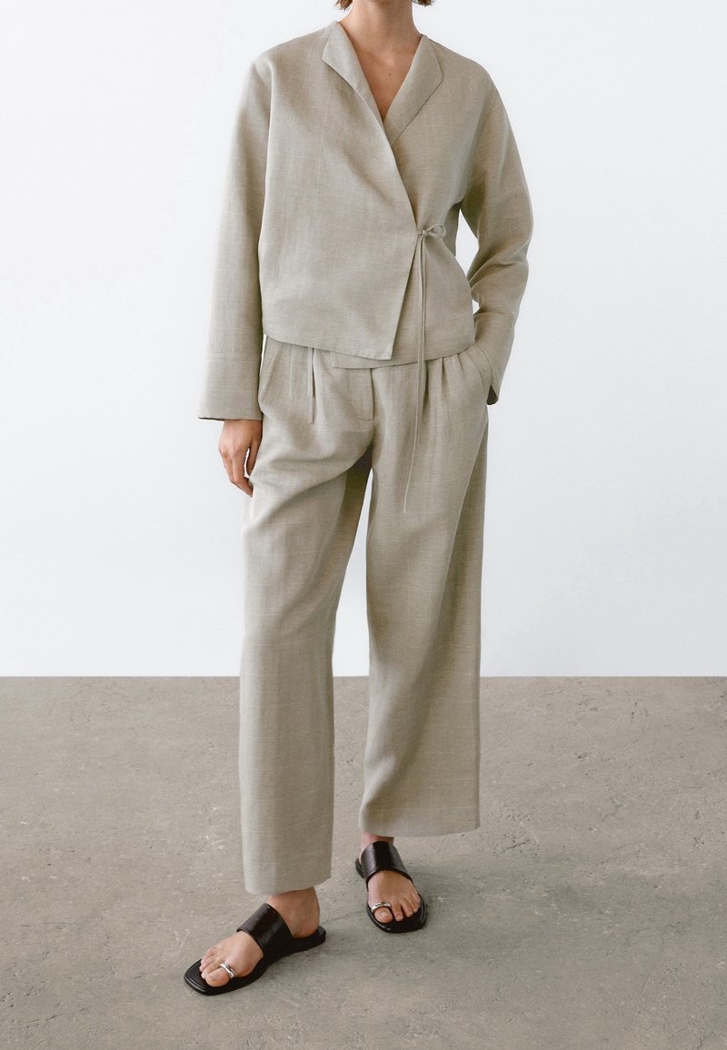 Personne portant une veste longue beige style cache-cœur avec manches longues, un pantalon large assorti et des sandales noires ouvertes avec anneaux d'orteils.