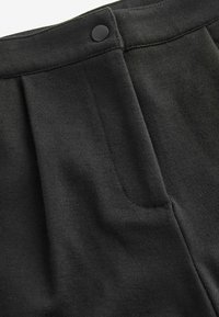 Zwarte stoffen broek met discrete voorplooien, een zijzak en een zwarte knoop op de tailleband. Gladde textuur en slank ontwerp.
