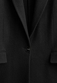 Manteau en laine noire avec une fermeture à un seul bouton noir, des revers crantés et des poches à rabat visibles sur les côtés.