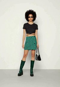 Mulher com cabelo afro, usando um top preto curto, saia floral verde, botas verdes até ao joelho, óculos de sol, segurando uma mala preta, de pé contra uma parede lisa.