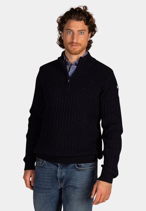Marineblauwe ribgebreide trui met een ritskraag, voorzien van kleurspetters en een strakke pasvorm. Draagt over een geknoopte, patroonshirt.