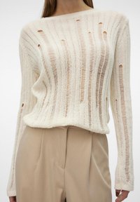Pull crème avec des motifs rayés verticaux et des découpes. Manches longues et coupe décontractée. Associé à un pantalon beige.