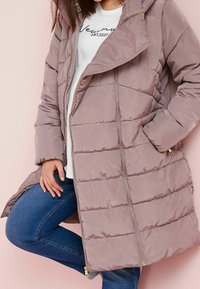 Woman wearing a mauve puffer coat over a white printed shirt and blue jeans, posing with one leg raised against a pale pink background.