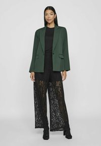 Grønn blazer med skreddersydd design, kombinert med svarte blonder vide bukser med blomster mønster og gjennomsiktige paneler. Svarte ankelboots.