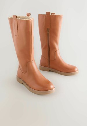 Bottes en cuir marron hauteur genou avec une fermeture éclair sur le côté et une languette pour tirer. Doublure intérieure douce et semelle plate en caoutchouc tan avec des détails cousus.