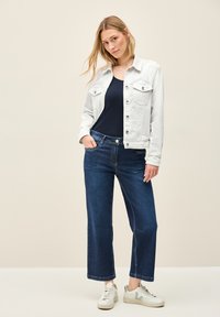 Vit denimjacka med fickor, marinblå topp och vida mörkblå jeans. Ljust färgade sneakers med grå accenter fulländar outfiten.