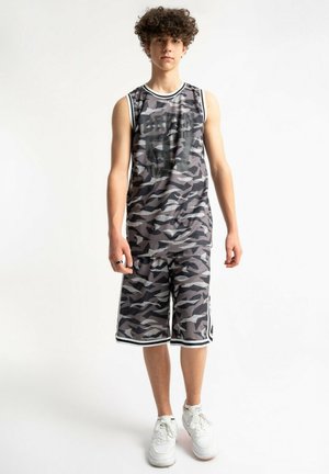 Nastolatek stoi ubrany w czarną i szarą kamuflażową koszulkę na ramiączkach do koszykówki oraz spodenki, do tego białe trampki, na białym tle.