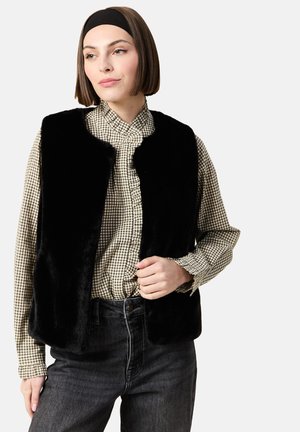 Gilet in pelliccia sintetica nera senza chiusura, indossato sopra una camicia button-up a fantasia. Il modello ha i capelli corti e una fascia nera.