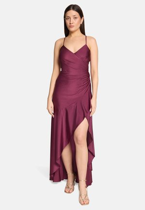 Vrouw staand, gekleed in een mouwloze, asymmetrische diep paarse jurk met dunne bandjes en gouden sandalen met bandjes, tegen een effen witte achtergrond.