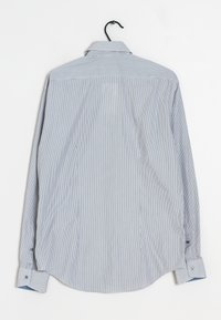 Chemise à rayures bleu clair et blanche à manches longues, col boutonné, avec des boutons aux poignets, suspendue à un crochet noir sur fond blanc.