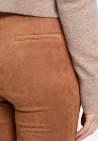 Pantalons en suède marron avec une texture lisse, comportant une poche arrière et une coupe ajustée. Le haut est un pull beige texturé.