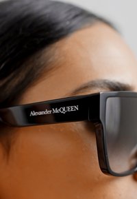Ochelari de soare supradimensionați, de culoare neagră, cu o ramă groasă și un finisaj mat. Logo-ul "Alexander McQueen" în alb pe lateral. Suprafață a lentilelor netedă.
