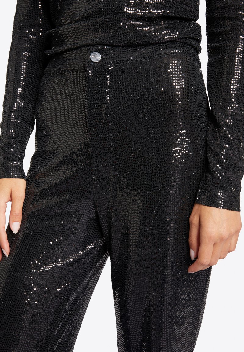 Czarna tkanina z cekinami o teksturowanym wzorze, dopasowany krój, z metalicznym guzikiem w detalach w talii. Widoczne długie rękawy.