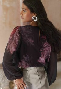 Durchsichtige schwarze Bluse mit rosa federartigen Mustern, Puffärmeln und einem Ausschnitt auf der Rückseite, kombiniert mit metallisch silbernen hoch geschnittenen Hosen.