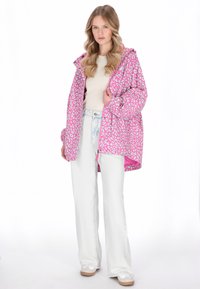 Rózsaszín leopárdmintás esőkabát kapucnival, könnyű, fényes textúrával. Fehér harangszárú farmerrel és sportcipővel párosítva.