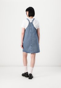 Denim kopējais kleitas ar neapstrādātu apakšmalu, regulējamām siksnām un brīvu piegriezumu. Nēsāta virs baltas T-krekliem, papildināta ar melnām kurpēm un zeķēm.