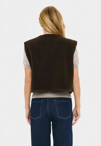 Persona rubia de espaldas, vistiendo un chaleco sin mangas de forro polar marrón oscuro sobre una camiseta de manga corta gris claro y jeans azul oscuro.