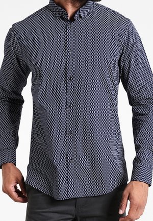 Homme portant une chemise bleu marine ajustée à boutons avec un petit motif géométrique blanc et un pantalon noir.