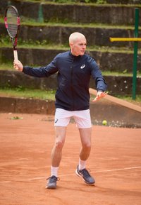 Tenisaš u tamnoplavoj zip-up jakni i bijelim šortsevima, drži reketa. Površina terena je od gline, s zelenim akcentima u pozadini.