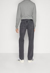 Jeans grigio scuro con taglio dritto, cinque tasche e un'etichetta del marchio visibile sulla cintura posteriore. Indossato con una maglietta grigia a maniche lunghe e sneakers.