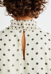 Blusa crema con pois neri; presenta un colletto smock e chiusura con bottoni sul retro; tessuto leggero e trasparente con una finitura testurizzata.