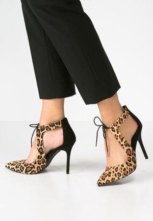 Pies con zapatos de tacón alto con estampado de leopardo y tiras negras para atar combinados con pantalones negros cortos sobre fondo blanco.