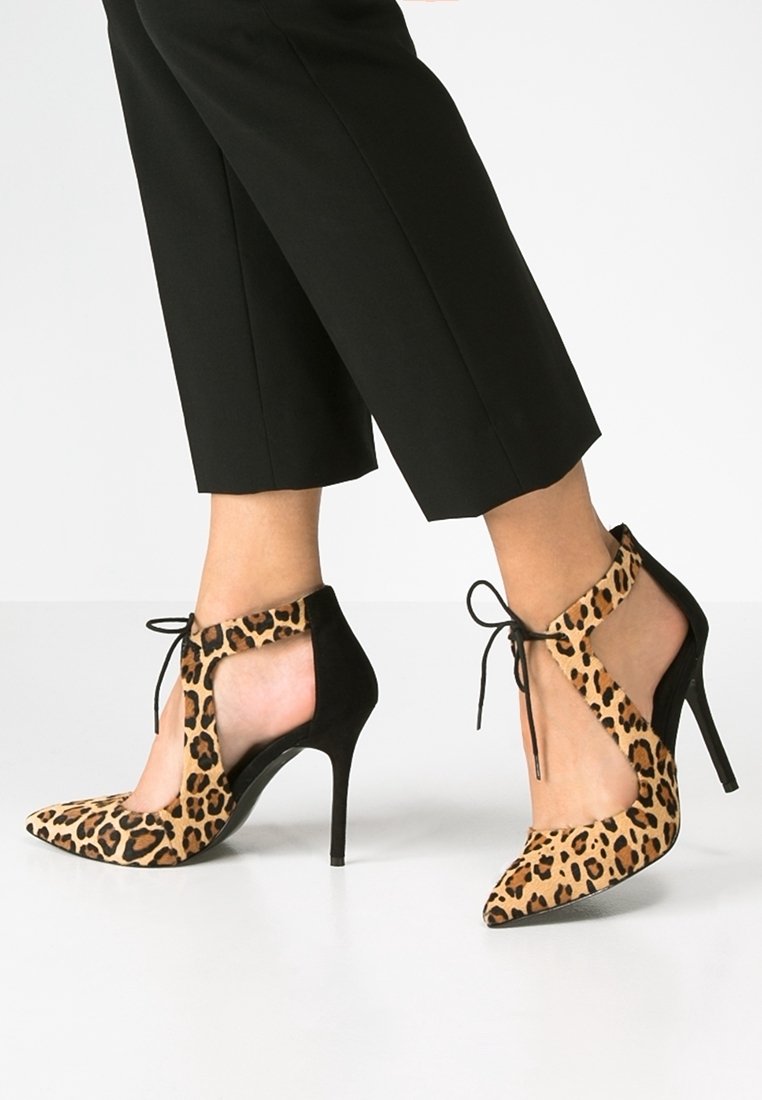 Pieds portant des chaussures à talons hauts imprimé léopard avec des lanières noires à lacets, associées à un pantalon court noir sur fond blanc.