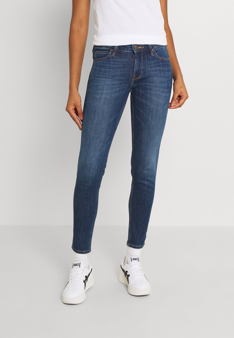 Person iført slim-fit blå jeans, hvide sokker og hvide Asics-sneakers, stående mod en ensfarvet lys baggrund.